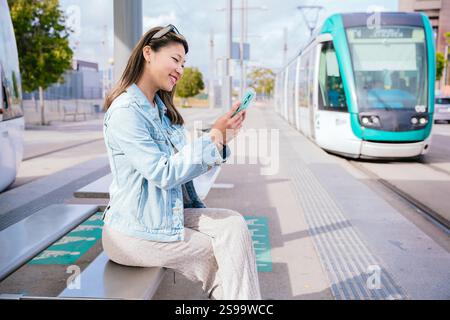 Smiling Young Woman Using Smartphone While Waiting for Tram at Station Stock Photo