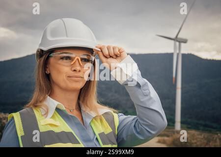 A female engineer is depicted wearing safety gear while working at a wind farm, showcasing her dedication to innovation in this beautiful landscape Stock Photo