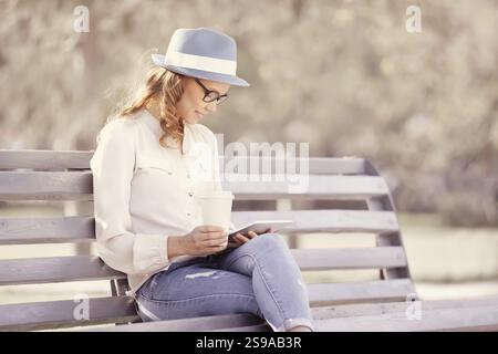 Happy young student with a tablet and a disposable coffee cup sitting on a bench reading in a summer park Stock Photo