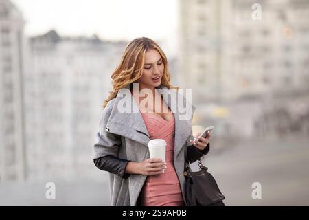 A businesswoman checking e-mails on her mobile phone and holding a coffee cup in her hand in front of an urban backdrop Stock Photo