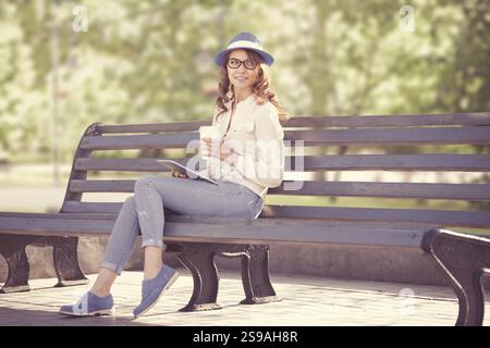 A happy young student with a tablet PC and a disposable coffee cup sits on a bench in a summer park Stock Photo