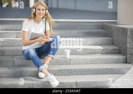 Happy young woman with vintage headphones and a cup of coffee to go, surfing the internet on a tablet PC, listening to music and sitting on a staircas Stock Photo
