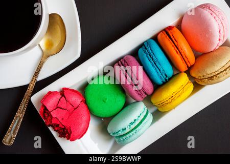 A white cup of coffee and multicolored macaroons on a white plate on a black background. French traditions Stock Photo