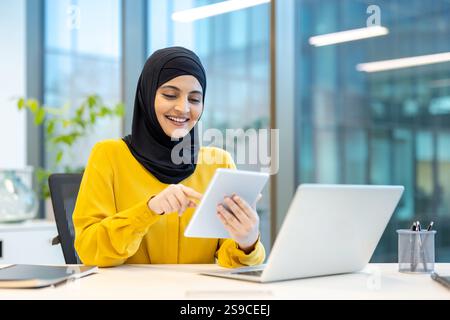 Successful woman in hijab using app on tablet computer. Arab office worker smiling contentedly, working inside office at workplace. Stock Photo