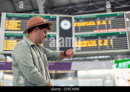 Young latin male tourist wearing a hat, checking the time on a smartwatch while waiting for train departure, glancing at the timetable display in the Stock Photo