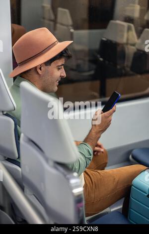 vertical Young Latin tourist wearing a stylish hat, using a mobile phone while enjoying a train journey, sitting next to his suitcase and embracing th Stock Photo