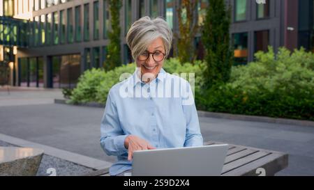 Happy middle-aged woman Caucasian mature senior business female talk online using laptop device outdoors adult cheerful businesswoman chatting with Stock Photo