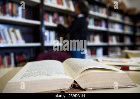 An open book lies on a table in a library, with bookshelves and a reader in the background. Stock Photo