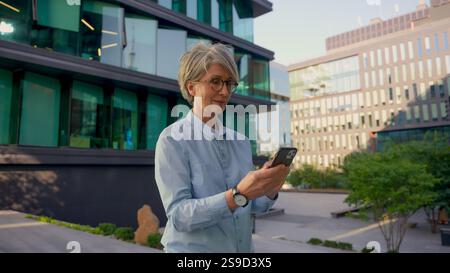 Mature Caucasian businesswoman smiling business lady entrepreneur employer middle-aged thoughtful woman using mobile phone outdoors browsing Stock Photo