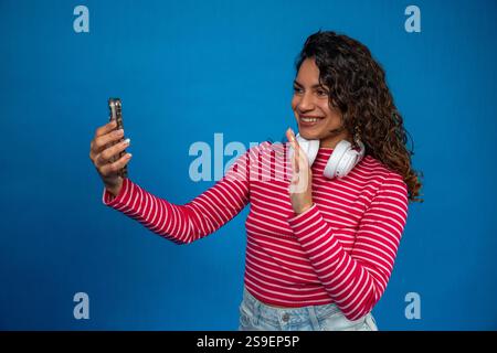 Young smiling woman with headphones making a video call with her phone waving with her hand on a blue background Stock Photo
