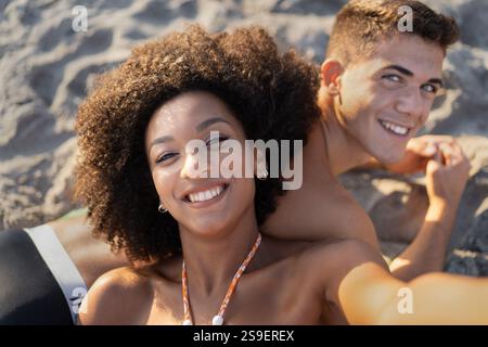 Cheerful multiracial couple taking a selfie on a sandy beach during a sunny summer day, capturing a joyful and carefree moment. Stock Photo