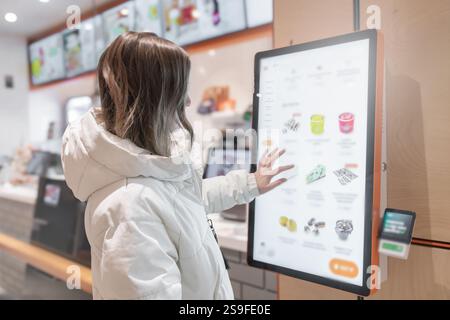 A 30 -35-year-old woman in a white winter jacket orders food through the screen menu at a fast food restaurant . A young individual is actively using Stock Photo