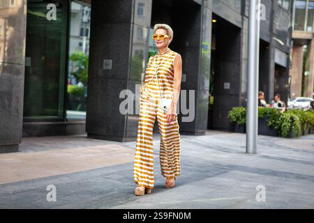 Fashionable Mature Woman Walking on City Street in Stylish Striped Yellow Suit with Pants and Top, Handbag, Wearing Heeled Shoes and Sunglasses. Elega Stock Photo