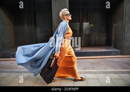 Stylish Woman Walking City Street In Fashionable Clothes, Wearing Long Beige Silk Dress, Blue Cape Coat, Carrying Large Black Handbag, Sunglasses And Stock Photo