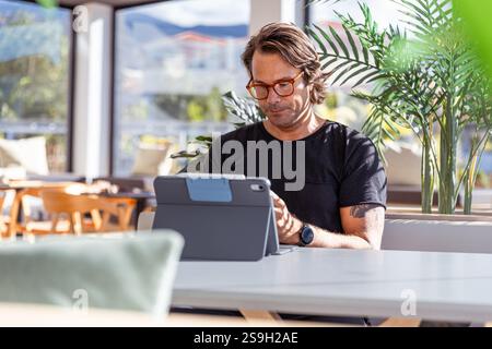 A man is sitting at a table working with his tablet and looking serious and concentrated. Stock Photo