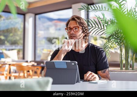 A man is sitting at a table in an office with a tablet working with a pensive expression Stock Photo