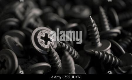 A detailed macro shot of black drywall screws with cross-shaped heads arranged in a dense pile. Monochromatic image suitable for industrial or mechani Stock Photo