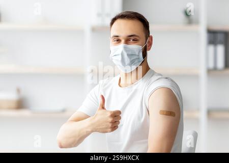 Young guy in face mask having plaster bandage on his arm after getting covid-19 vaccine injection, showing thumb up Stock Photo