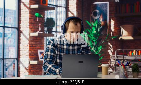 Employee completing spreadsheets on laptop and listening music. Worker in home office wearing headphones editing documents on notebook, updating reports and files, camera B Stock Photo
