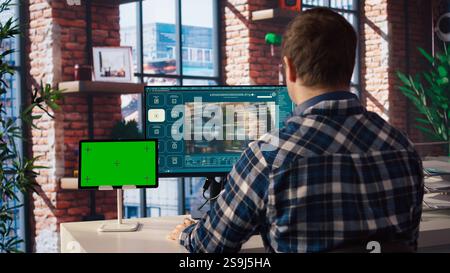 Man in home office uses AI program turning text prompt into videos next to isolated screen tablet. Person using text to video generative models on computer and chroma key device, camera A Stock Photo
