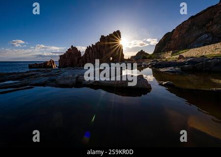 The sun is rising behind the blood-red and grey porphyry peaks of Arbatax, Rocce Rosse, reflected in a small pond. Arbatax Sardinia Italy FB 2024 1355 Stock Photo