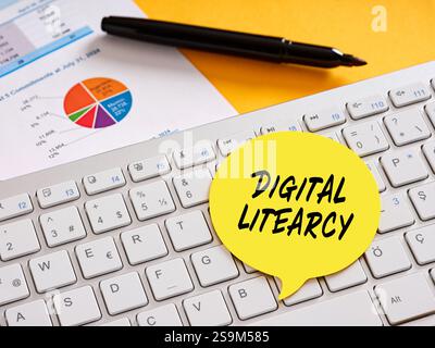 Yellow sticky note labeled digital literacy resting on a computer keyboard, surrounded by charts and a pen, representing online education Stock Photo