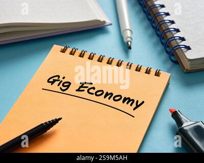 Orange notebook with gig economy written on it, accompanied by markers and notepad on a blue surface, representing modern work trends Stock Photo