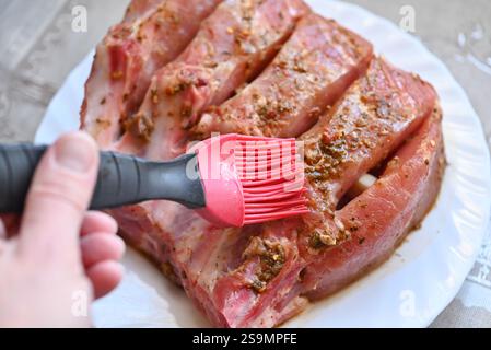 Close-up view of marinating raw pork ribs with a red silicone brush on a white plate, creating an appetizing scene. Isolated on gray background with a Stock Photo