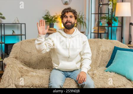 POV of Indian young man looking at camera, making video webcam conference call with friends or family, enjoying conversation at home room apartment. Guy with curly hair sits on sofa waving hello. Stock Photo