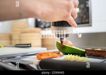 Chef drizzling soy sauce over grilled salmon with avocado slices and garnish, highlighting food preparation in a professional Stock Photo