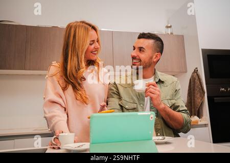 Happy Couple Enjoying a Wonderful Morning with Coffee and Laughter in a Cozy Kitchen While Engaging in Lighthearted Conversation and Sharing Joyful Moments Together with a Tablet Set on the Counter. High quality photo Stock Photo