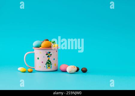 Colorful Easter chocolate eggs in pink enamel mug against blue background. Minimal Easter concept. Creative festive spring holiday layout. Stock Photo