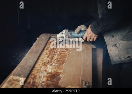 Person using an angle grinder on a wooden surface in a dimly lit workshop Stock Photo