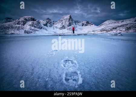 Scandinavian winter Landscape of Snowy Mountain peaks and sea coast, frozen lake water with footsteps in snow and photographer person with camera. Lof Stock Photo
