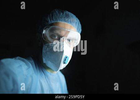 Doctor wearing goggles and mask in blue scrubs with a dark background Stock Photo