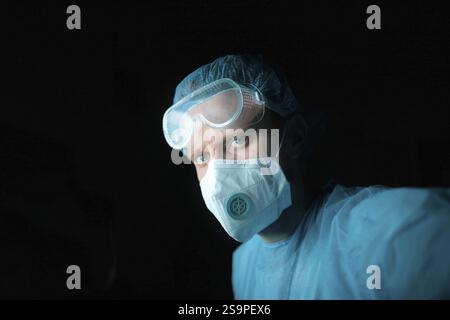 Doctor in blue scrubs wearing goggles and face mask, set against a dark backdrop Stock Photo