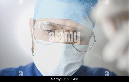 Close-up of a male doctor wearing glasses, a surgical mask, and blue scrubs, focused on a task Stock Photo