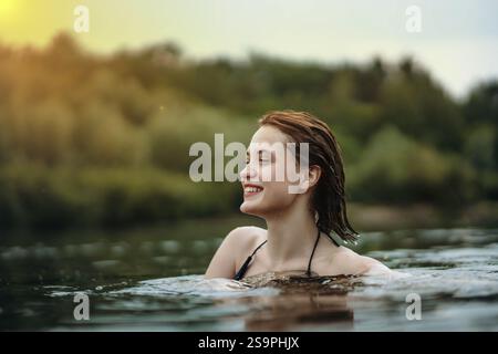A content woman smiles while relaxing in water, surrounded by forest at sunset Stock Photo