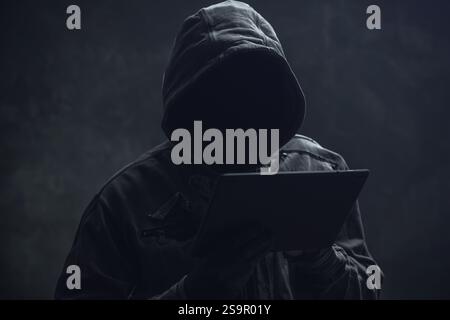Hooded computer hacker using digital tablet, selective focus Stock Photo