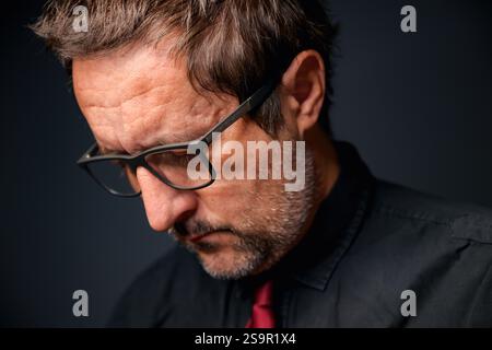 Portrait of serious concerned businessman entrepreneur wearing eyeglasses and black shirt in dark office interior, closeup low key with selective focu Stock Photo