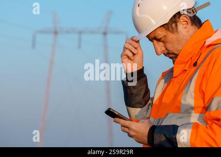 Electrical engineer using smartphone out in the field close to electricity pylons, selective focus Stock Photo