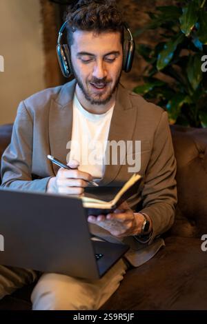 Focused man wearing headphones, writing notes in a notebook while working on a laptop in a cozy indoor environment, ideal for remote work or creative Stock Photo