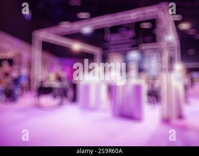 A blurry image of an event booth setup under pink and purple lighting, featuring modern structures and indistinct attendees in a dynamic indoor venue. Stock Photo