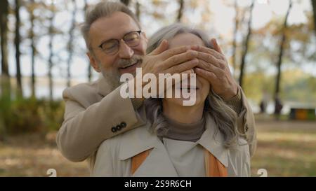 Mature senior retired Caucasian man affectionate touch closing woman eyes with hands surprise in city romantic meeting date in park smiling happy old Stock Photo