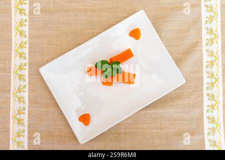 A creative arrangement of sliced orange carrots on a white plate, garnished with fresh mint leaves. The plate is set against a beige tablecloth with d Stock Photo