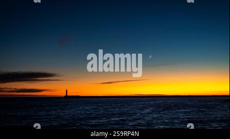 A serene sunset over the ocean, featuring a lighthouse silhouette against a colorful sky transitioning from orange to deep blue, with a crescent moon Stock Photo