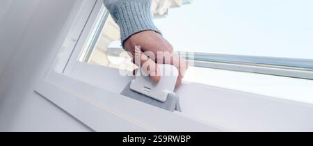 a man cleans the grooves of a white sliding window frame using a cleaning device, in a panoramic format to use as web banner Stock Photo
