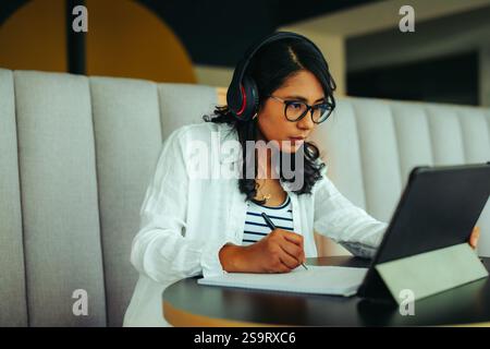 A college student is seen intently working on an assignment, using a tablet and taking notes while wearing headphones in a comfortable study space. Stock Photo