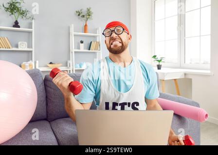 Serious Man Exercising With Dumbbells At Home Using Laptop In Funny Outfit Stock Photo