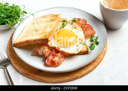 Breakfast sandwich  with fried egg, bacon and green pea sprouts served on plate close up. Fried sunny side egg, bacon on toast with microgreens and mo Stock Photo